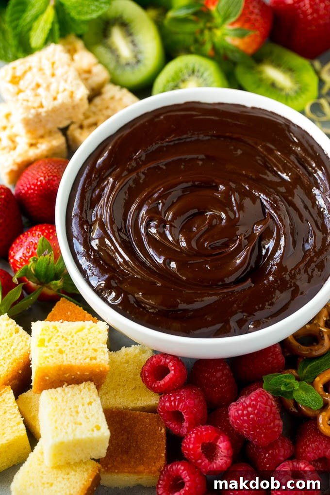 A bowl of chocolate fondue surrounded by an array of dippers including fruit, cake, rice krispy treats, and pretzels.