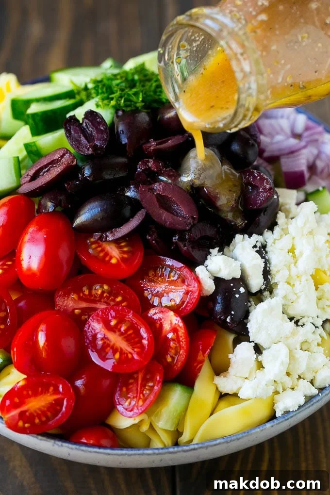 A generous stream of homemade garlic and herb dressing being poured over a bountiful bowl of Greek tortellini and colorful vegetables, ensuring every ingredient is perfectly coated.