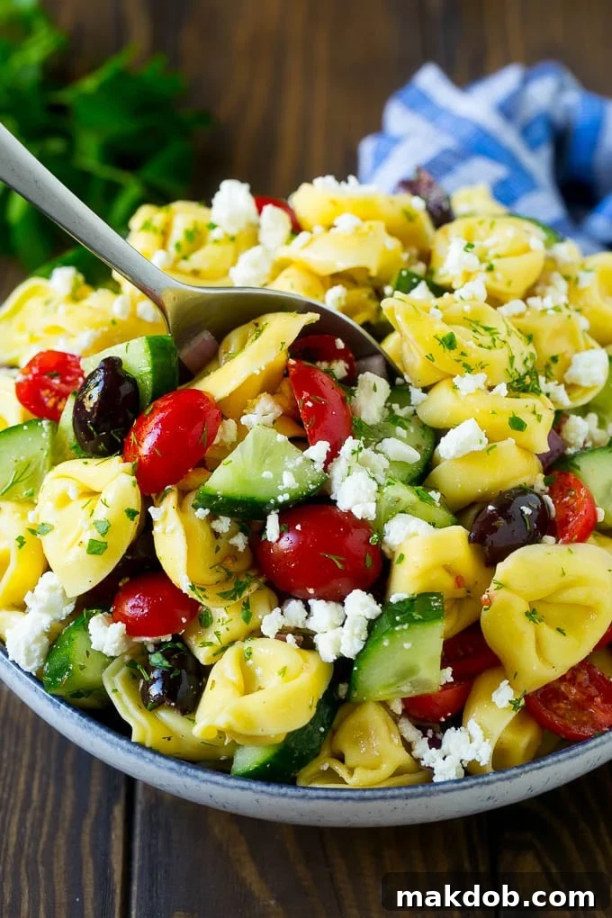 A large serving bowl filled with Greek tortellini salad, featuring a serving spoon ready to dish out the flavorful pasta and vegetable mixture.