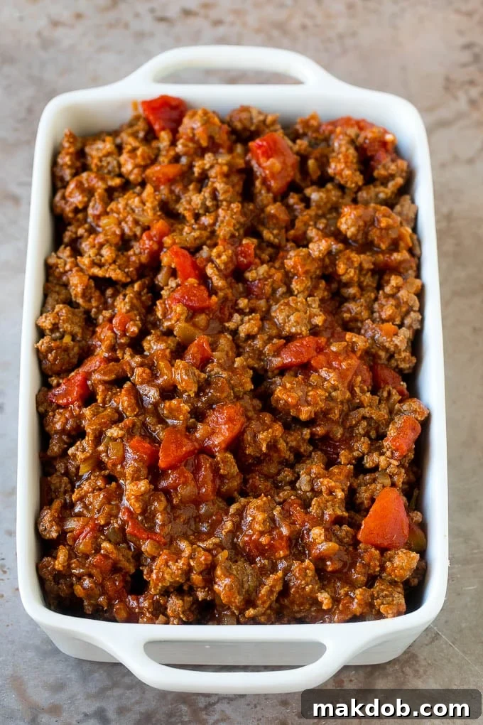 A savory cooked taco meat mixture, rich with spices, onions, and tomatoes, carefully spread as a layer in a casserole dish.