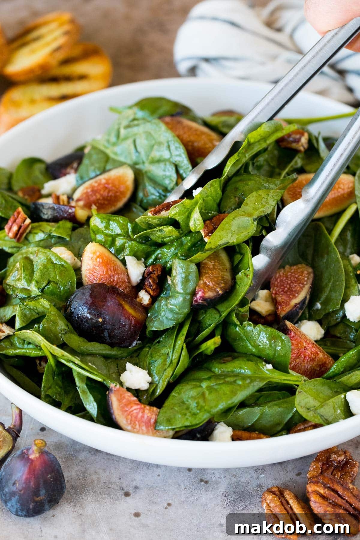 A pair of serving tongs lifting a portion of the fresh fig salad, highlighting its colorful ingredients and inviting presentation.