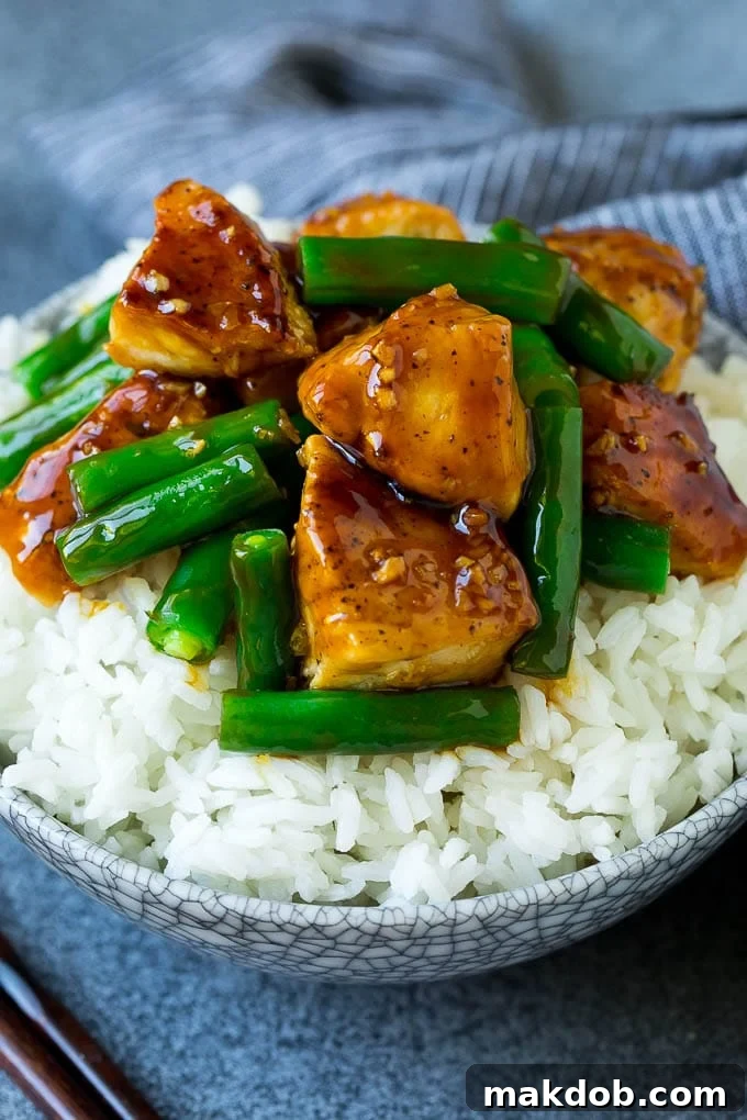 Honey garlic chicken and green bean stir fry served over white rice in a bowl, garnished with sesame seeds.