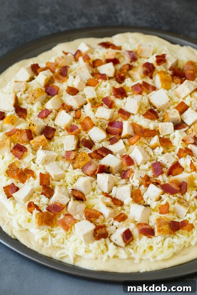 A pizza crust topped with rich Alfredo sauce, shredded mozzarella cheese, cooked chicken, and crispy bacon, ready for baking.