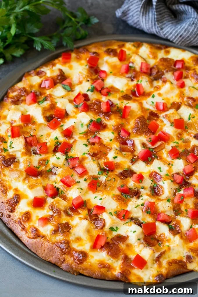 Chicken Alfredo Pizza Recipe #pizza #chicken #bacon #dinner #dinneratthezoo A whole Chicken Alfredo Pizza topped generously with white sauce, melted mozzarella cheese, tender chicken pieces, crispy bacon, and fresh diced tomatoes, with a scattering of green parsley, ready to be sliced.