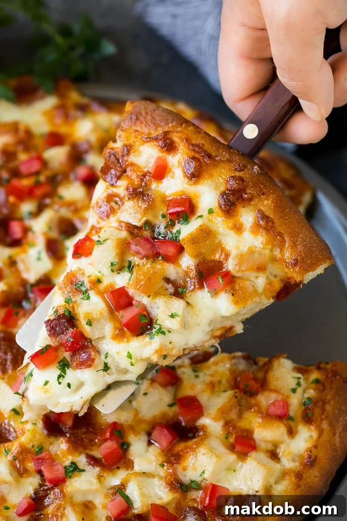 Chicken Alfredo Pizza Recipe #pizza #chicken #bacon #dinner #dinneratthezoo A hand holding a pizza spatula with a large, perfect slice of Chicken Alfredo Pizza, showcasing the creamy sauce, melted cheese, chicken, bacon, fresh tomatoes, and parsley against a blurred background of the whole pizza.