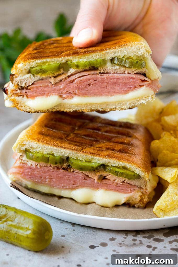 Sliced Cuban Sandwich A hand holding up half of a perfectly pressed Cuban sandwich, showcasing the cross-section of crispy bread, melted cheese, ham, pork, and pickles.