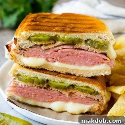 A perfectly pressed Cuban sandwich, golden brown and oozing with melted Swiss cheese, layered with ham, roasted pork, and pickles, sliced diagonally to reveal its delicious layers.