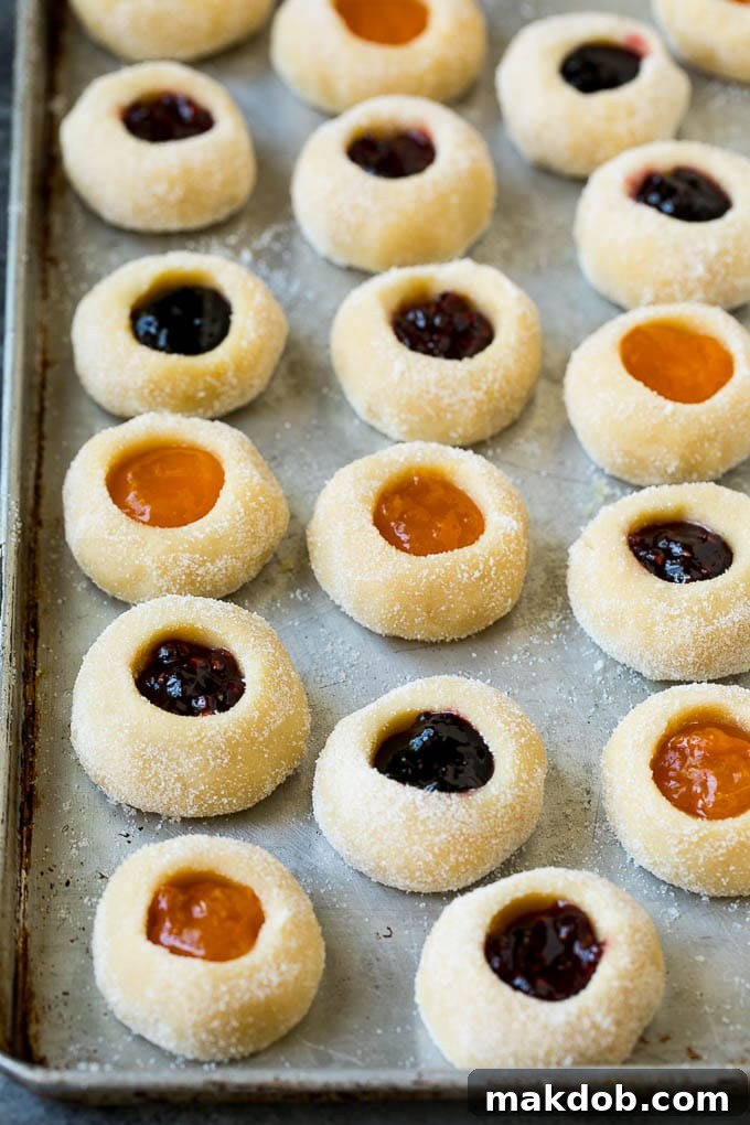 Classic Jam Thumbprint Cookies | Sugar Cookies with Jam Filling #cookies #baking #christmas #dessert #homemade Close-up view of raw cookie dough balls on a baking sheet, each carefully filled with vibrant fruit jam, prior to baking.