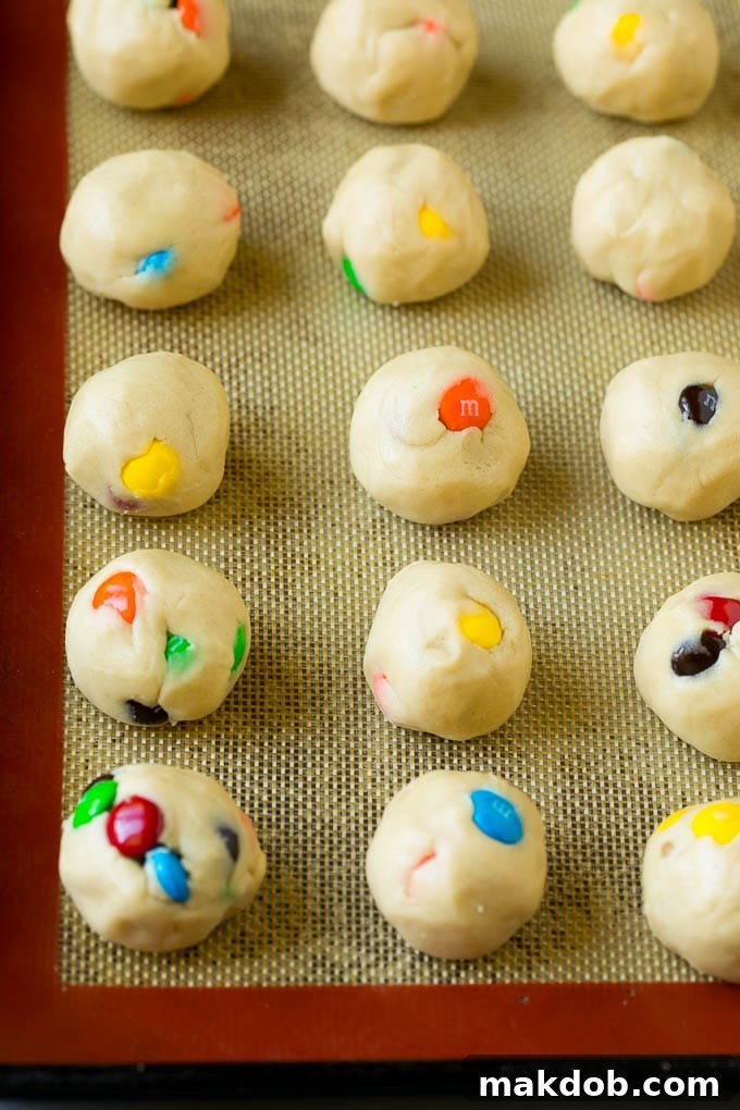 Uniformly sized balls of M&M cookie dough, adorned with extra candies, neatly arranged on a baking sheet, ready for chilling and baking.