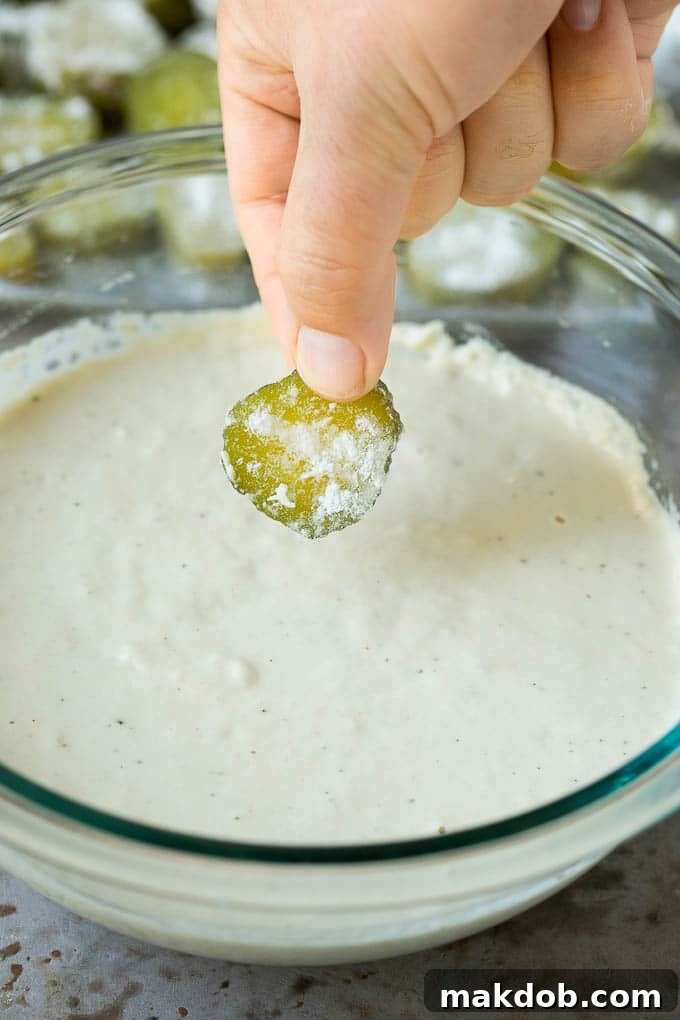 A pickle slice being expertly dipped into a bowl of light, seasoned beer batter, ensuring full coverage for an ultimately crispy fry.