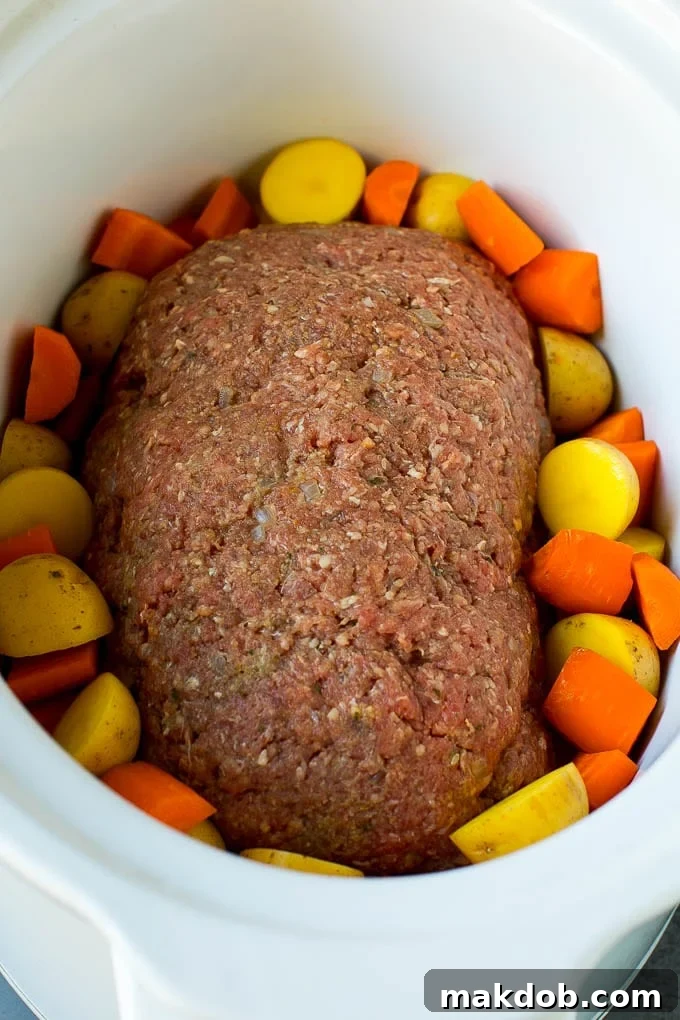 Ground beef shaped into a loaf in a crockpot, surrounded by carrots and potatoes before cooking.