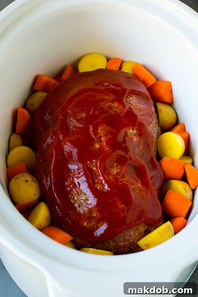 A loaf of ground beef topped with ketchup in a slow cooker, surrounded by carrots and potatoes.