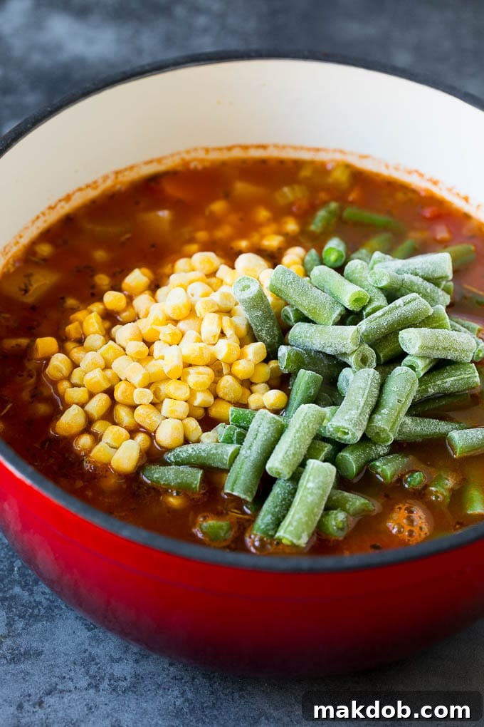 A close-up shot of steaming vegetable soup in a pot, with brightly colored frozen corn and crisp green beans sprinkled on top, having just been added.