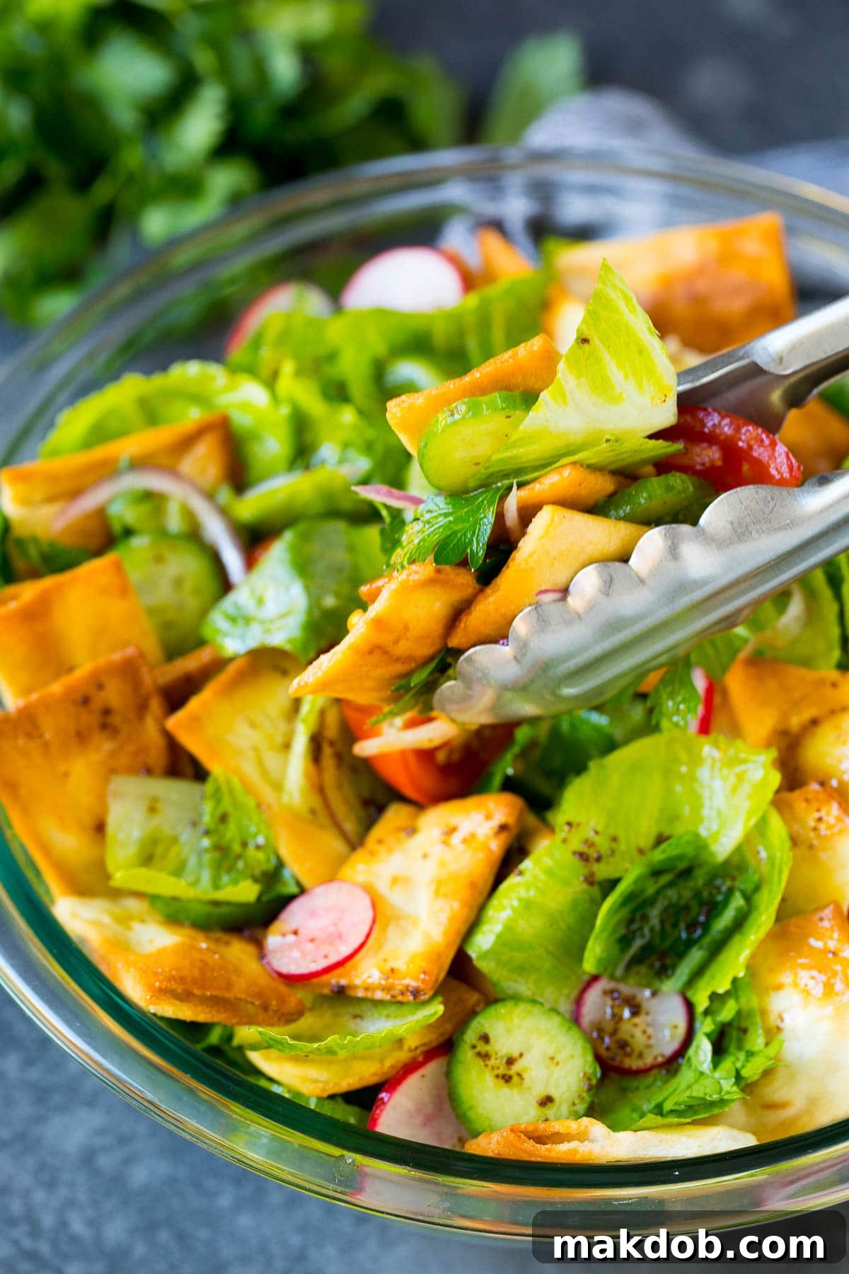 Tongs gracefully serving a fresh portion of Fattoush salad from a large bowl, highlighting its crisp textures and vibrant colors.