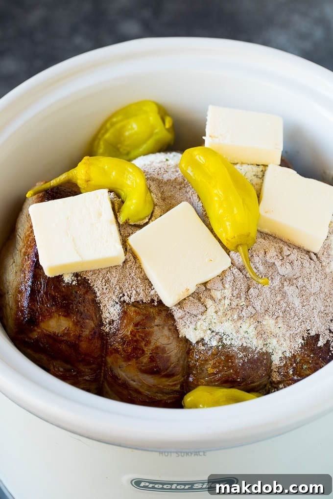 Seasoning Mississippi Pot Roast in Slow Cooker A pot roast in a slow cooker, generously topped with dry seasoning packets, butter slices, and whole pepperoncini peppers.