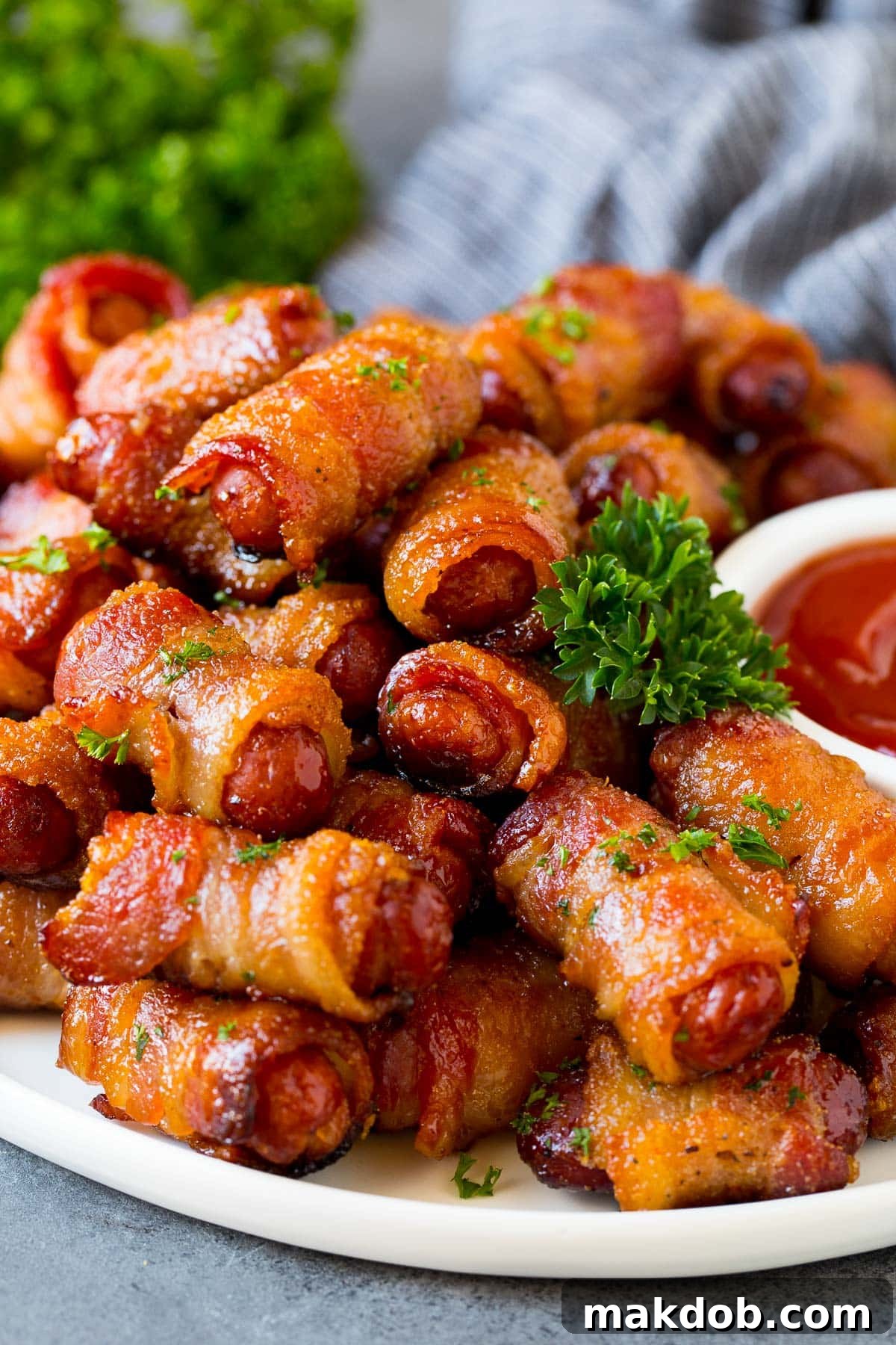 A serving plate of perfectly baked bacon wrapped smokies garnished with fresh parsley, with a side of ketchup for dipping.