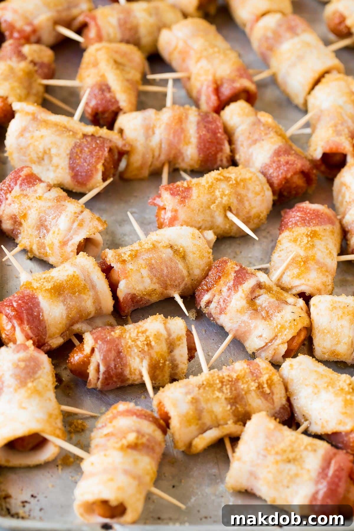 Close-up shot of bacon wrapped smokies on a baking sheet, each secured with a toothpick, showing the caramelized brown sugar coating.