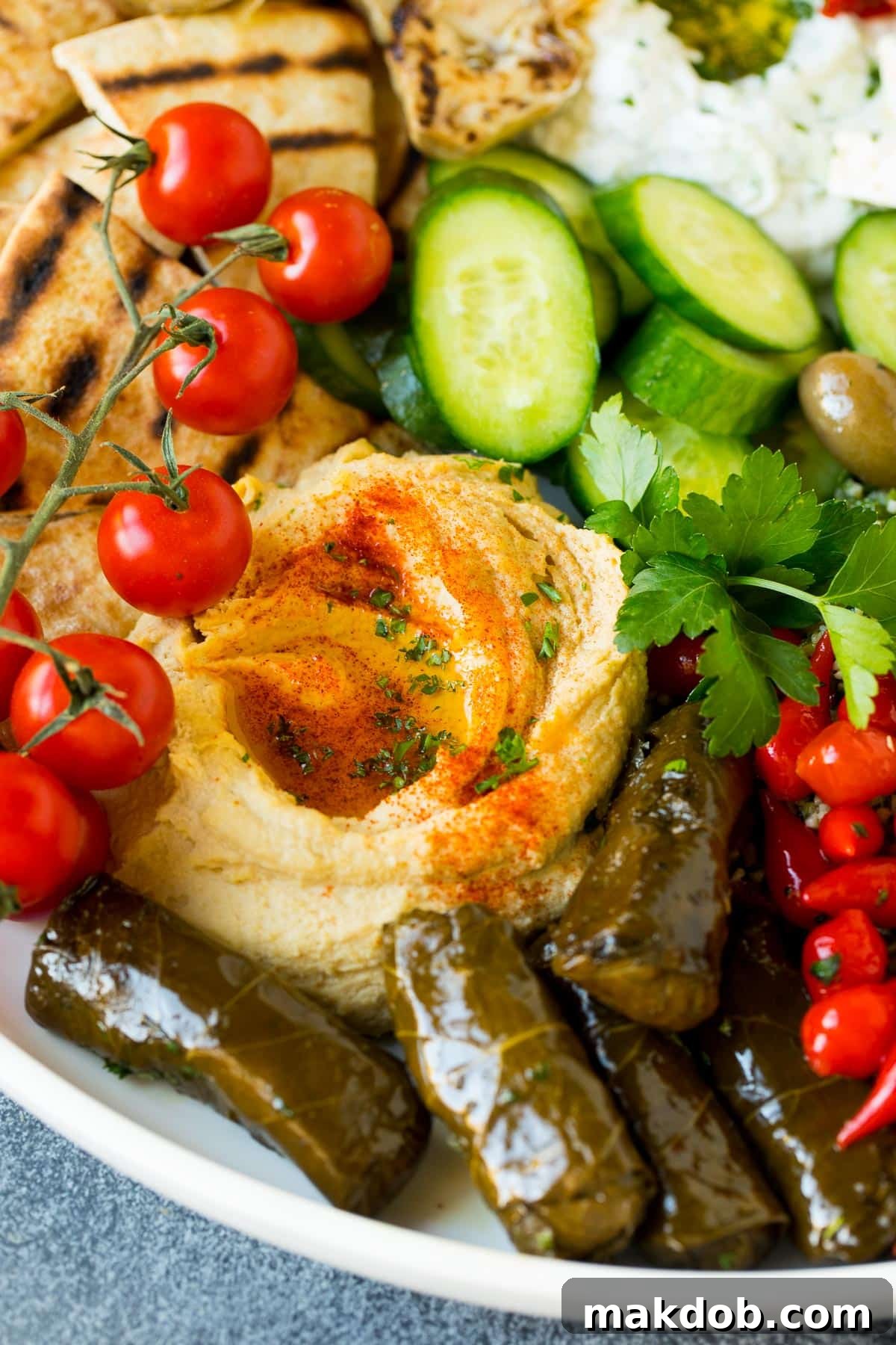 Homemade hummus served on a platter with fresh tomatoes, stuffed grape leaves, and crisp cucumbers, showcasing a delicious Mediterranean spread.