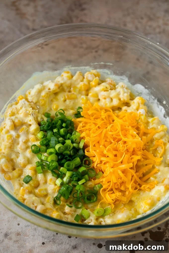 A vibrant bowl of corn fritter batter, generously topped with thinly sliced green onions and a mound of shredded cheddar cheese, ready for frying.
