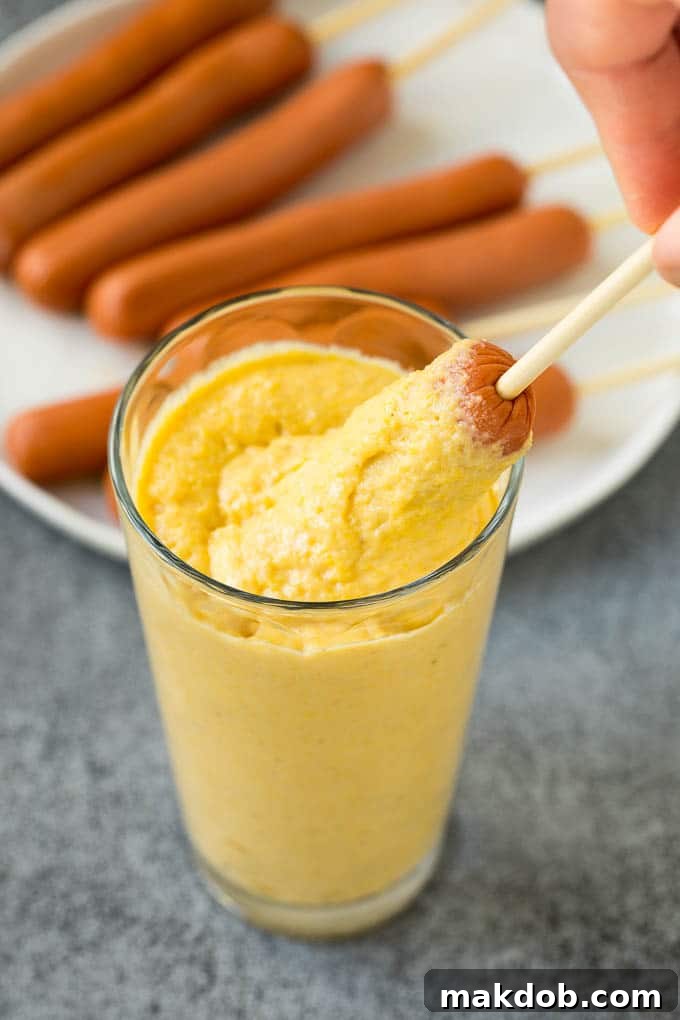 Homemade corn dogs are so easy to make and are even better than what you'd get at the fair! A hot dog on a stick being expertly dipped into a tall glass of rich corn batter, capturing the moment of preparation.