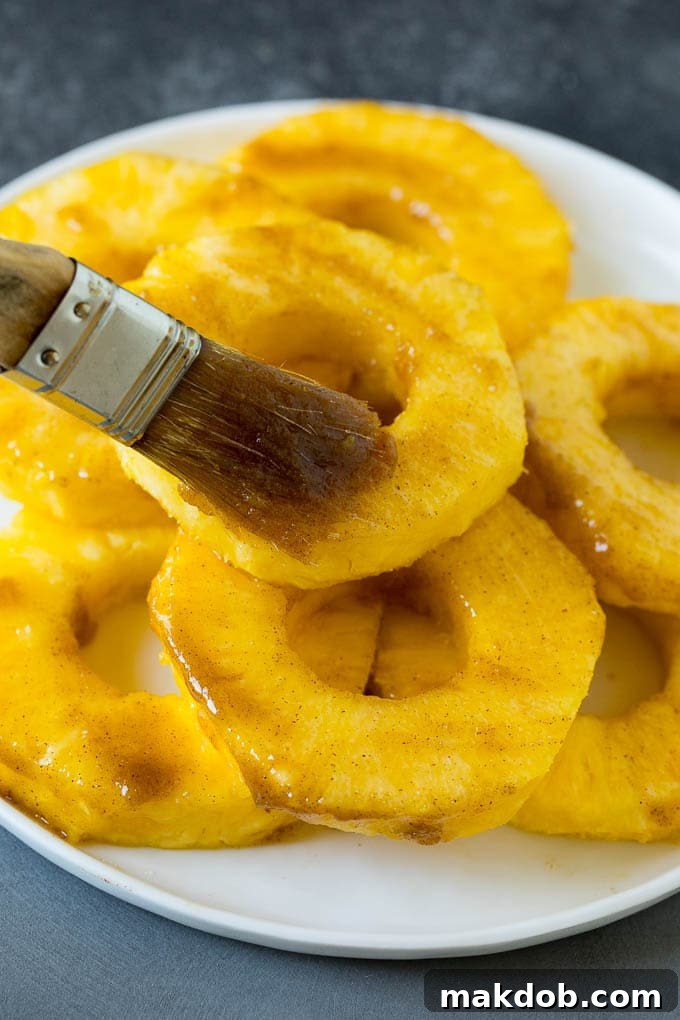 A basting brush coating fresh pineapple slices with a brown sugar and butter mixture before grilling.