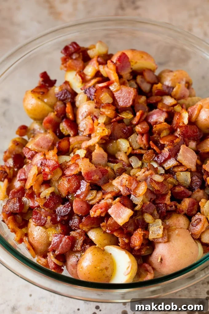 This German potato salad is boiled baby potatoes tossed in a warm bacon and mustard dressing, then garnished with fresh herbs. Cooked bacon and halved potatoes in a bowl.