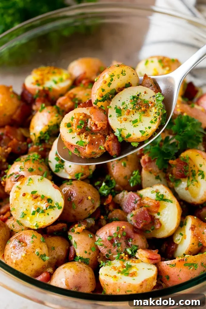 This German potato salad is boiled baby potatoes tossed in a warm bacon and mustard dressing, then garnished with fresh herbs. A spoon serving up a portion of German potato salad.