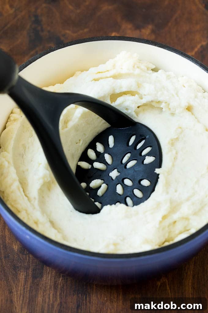 Mashing Potatoes with Cream Cheese A hand using a potato masher to blend boiled potatoes with butter, milk, cream cheese, and seasonings in a large pot.