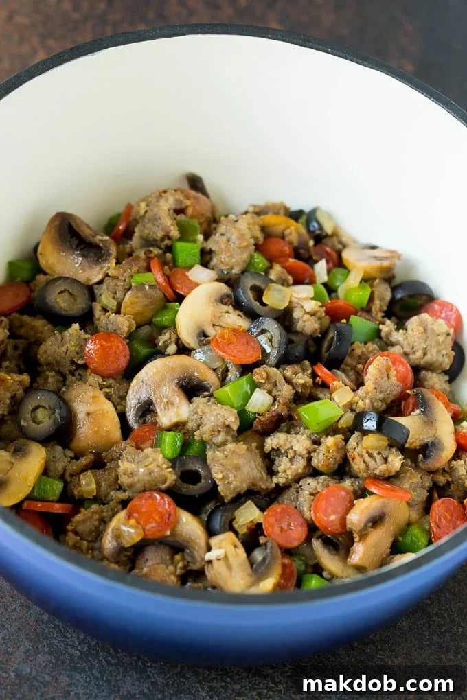 Sausage, mushrooms, olives, pepperoni and peppers cooking in a pot for the casserole base. A large pot on a stovetop, filled with browned Italian sausage, diced green bell peppers, sliced mushrooms, black olives, and mini pepperoni, mixed and ready for the sauce and pasta to be added.