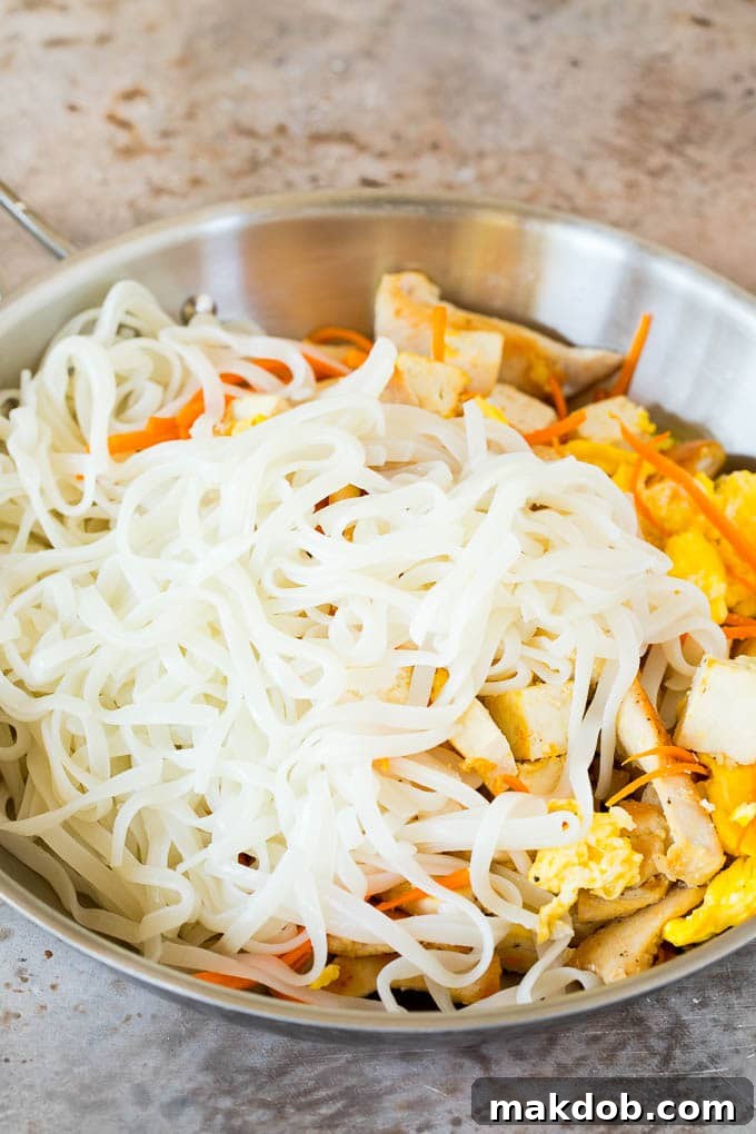 Stir-fried rice noodles in a pan, mixed with scrambled eggs, shredded carrots, chicken, and tofu, showing the vibrant colors of the dish.