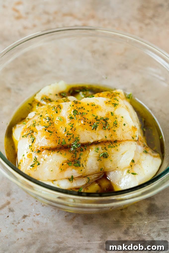 This baked cod is marinated with olive oil, garlic and fresh herbs, then roasted to tender and flaky perfection. Fish fillets in a bowl of marinade.