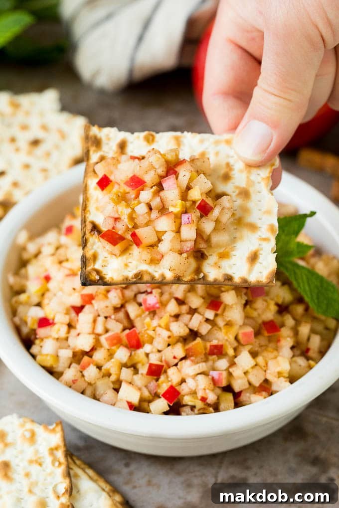 This charoset recipe is a blend of apples, walnuts and honey, all mixed together to make a sweet spread that's an essential part of any Passover meal. A hand holding a piece of matzo topped with a generous serving of homemade charoset.