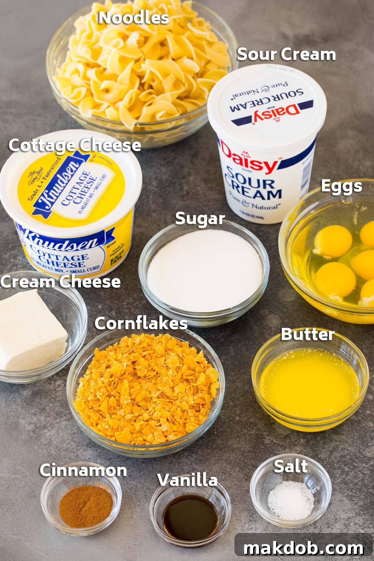 Golden Noodle Delight 3 Individual bowls displaying all the ingredients required for noodle kugel, including dry egg noodles, various dairy products like sour cream and cottage cheese, eggs, granulated sugar, melted butter, cinnamon, vanilla extract, and a bowl of cornflakes.