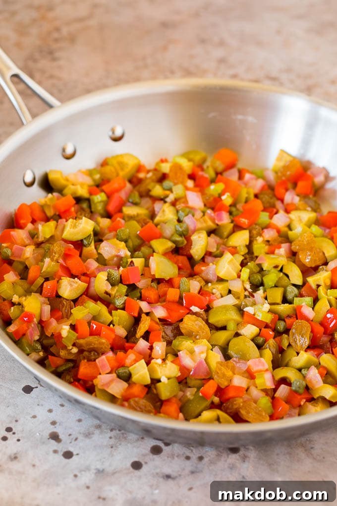 Vibrant mix of olives, sautéed red onions, bell peppers, celery, and capers simmering together in a cast-iron skillet, creating the aromatic base for a rich and authentic caponata.