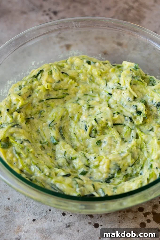 These zucchini fritters are crispy patties filled with shredded squash and flavored with garlic and herbs. Batter to make zucchini pancakes in a mixing bowl.
