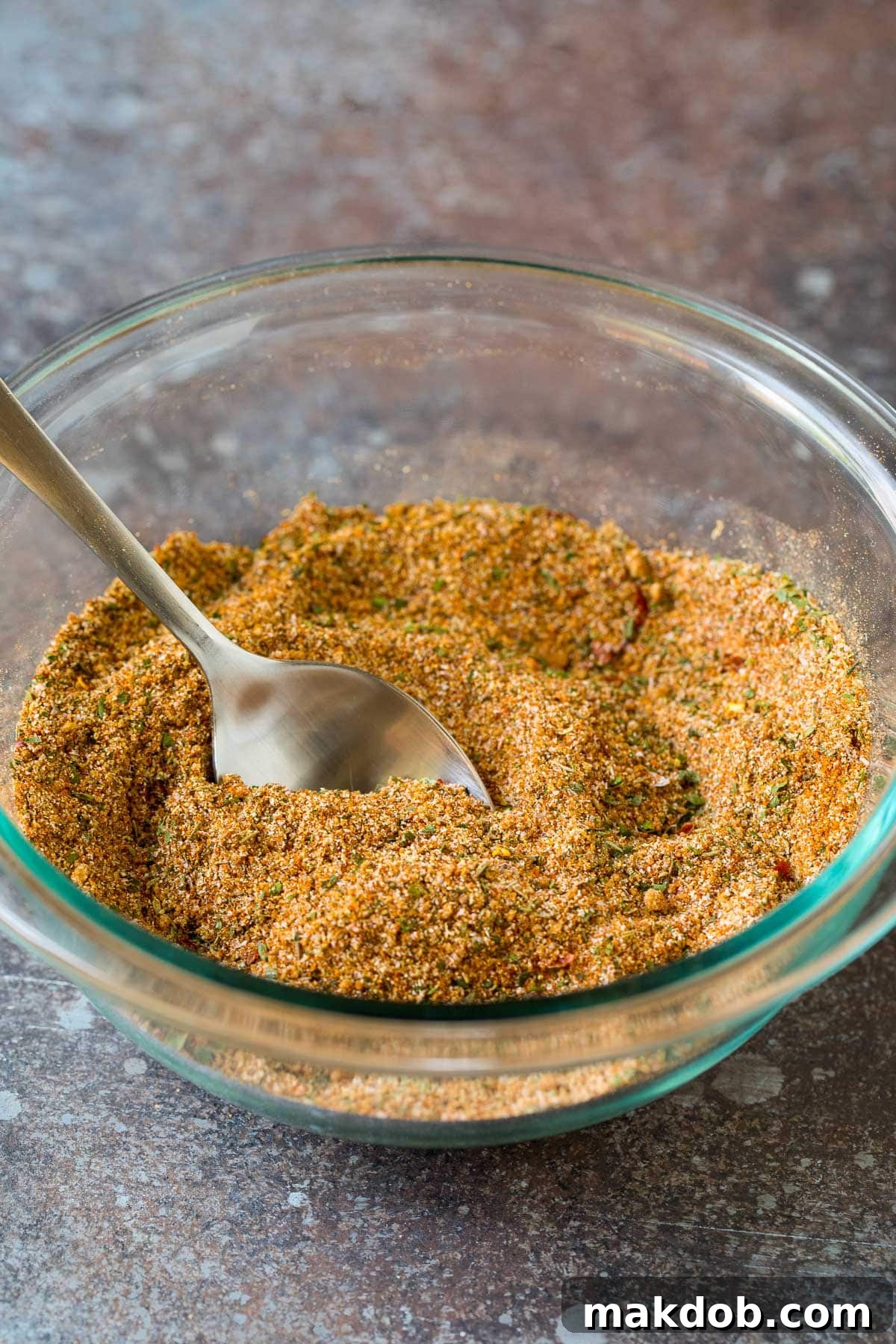A rustic bowl filled with rich, brown homemade jerk spice mix, with a small wooden spoon resting inside, capturing its inviting aroma and texture.