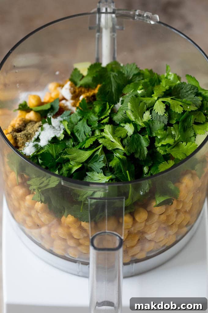 Soaked dried chickpeas, fresh herbs like parsley and cilantro, and an array of spices like cumin and coriander, arranged in a food processor, ready for blending.