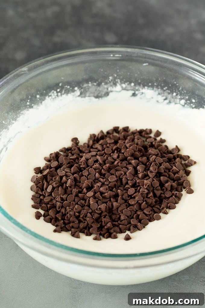 This cannoli dip is a blend of two types of sweet Italian cheeses, sugar and chocolate chips, all blended together to make a creamy dip that tastes just like the inside of a cannoli. A glass bowl filled with a creamy mixture of whipped ricotta and mascarpone cheese, topped with a generous sprinkle of miniature chocolate chips, ready for the final stir.