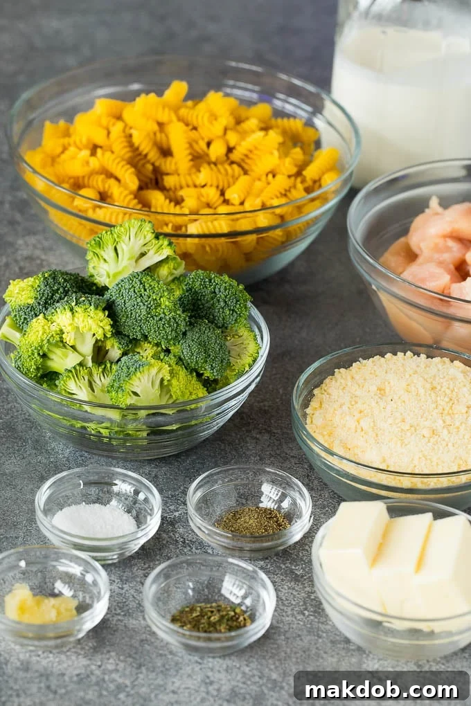 A vibrant collection of fresh ingredients laid out for chicken broccoli Alfredo, including uncooked rotini pasta, fresh broccoli florets, diced raw chicken, a stick of butter, and a bowl of grated Parmesan cheese.
