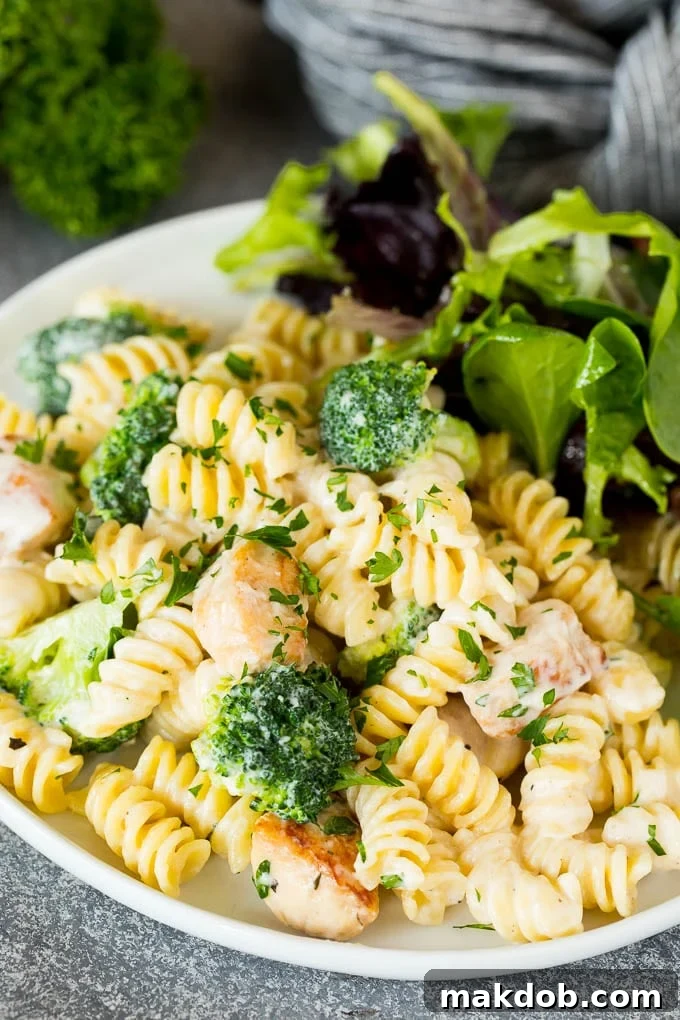 A close-up of a perfectly plated serving of chicken broccoli Alfredo pasta in a white bowl, accompanied by a fresh green side salad, ready for a delightful meal.