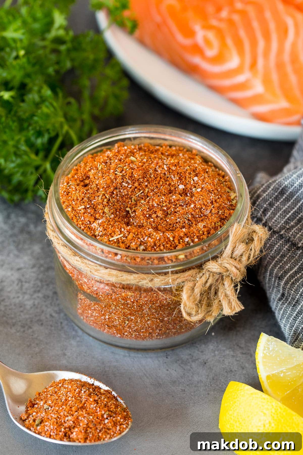 Signature Salmon Seasoning Blend 2 A jar of salmon seasoning with salmon, lemon wedges and parsley in the background.