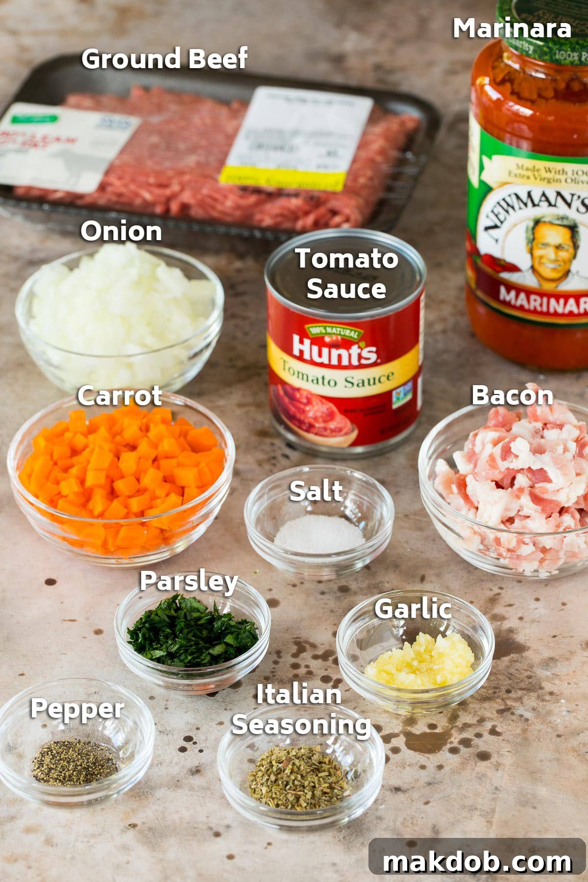 A collection of fresh ingredients laid out, including ground beef, strips of bacon, diced onions, carrots, minced garlic, marinara sauce, and canned tomato sauce.