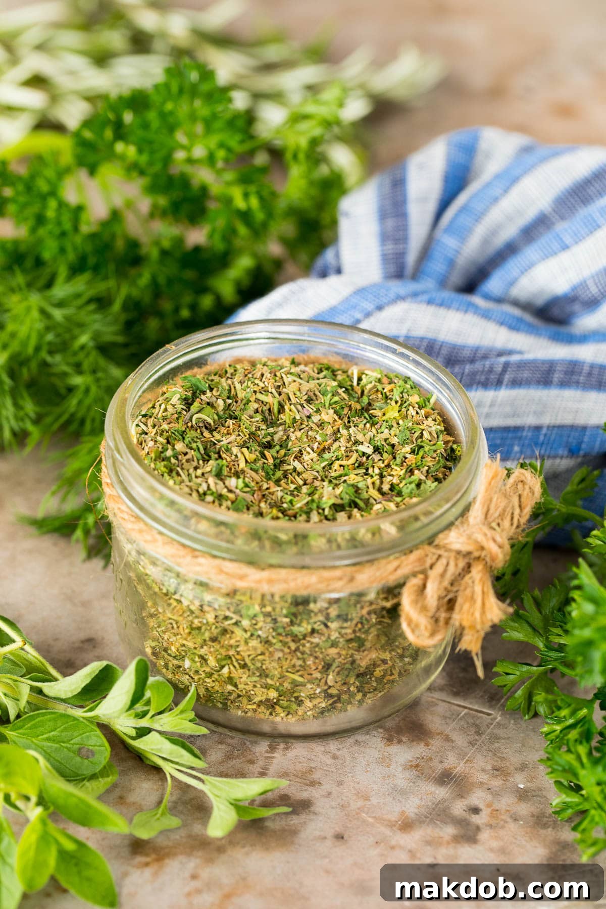 A jar of Greek Seasoning surrounded by fresh herbs, showcasing its natural ingredients.