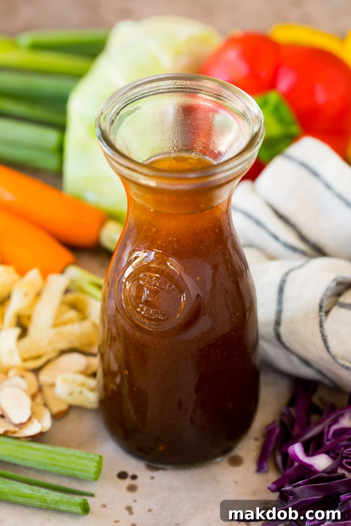 A beautifully presented jar of homemade Asian salad dressing, surrounded by fresh, colorful vegetables like cucumbers, carrots, and lettuce, suggesting its fresh ingredients and versatility.