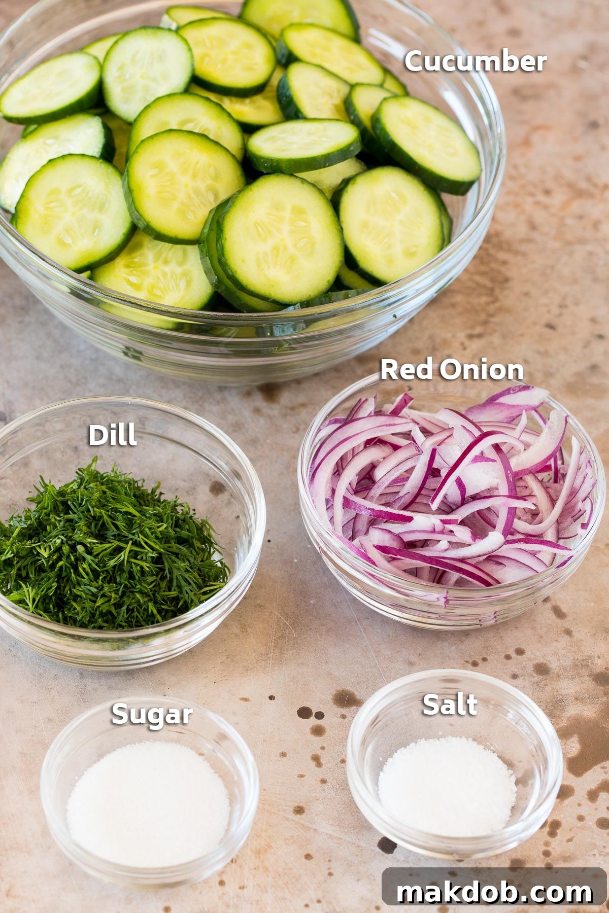 Bowls containing cucumbers, red onion, fresh dill, granulated sugar, and kosher salt, neatly arranged on a counter.