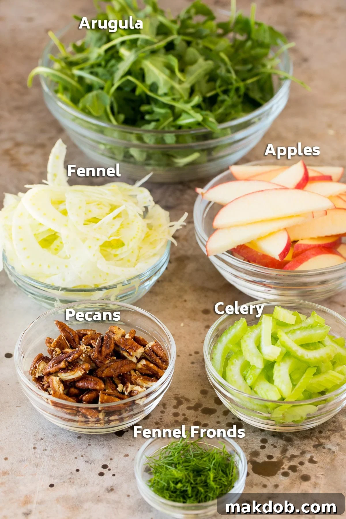 Individual bowls showcasing the fresh, key ingredients for fennel salad: vibrant arugula, thinly sliced apples, delicate fennel bulb slices, and crunchy candied pecans.