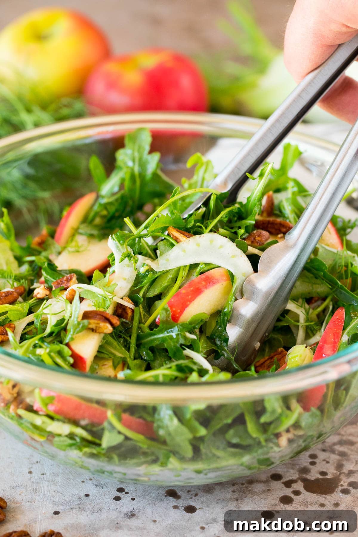 A pair of serving tongs lifting a generous portion of crisp fennel and apple salad from a large bowl, ready to be enjoyed.