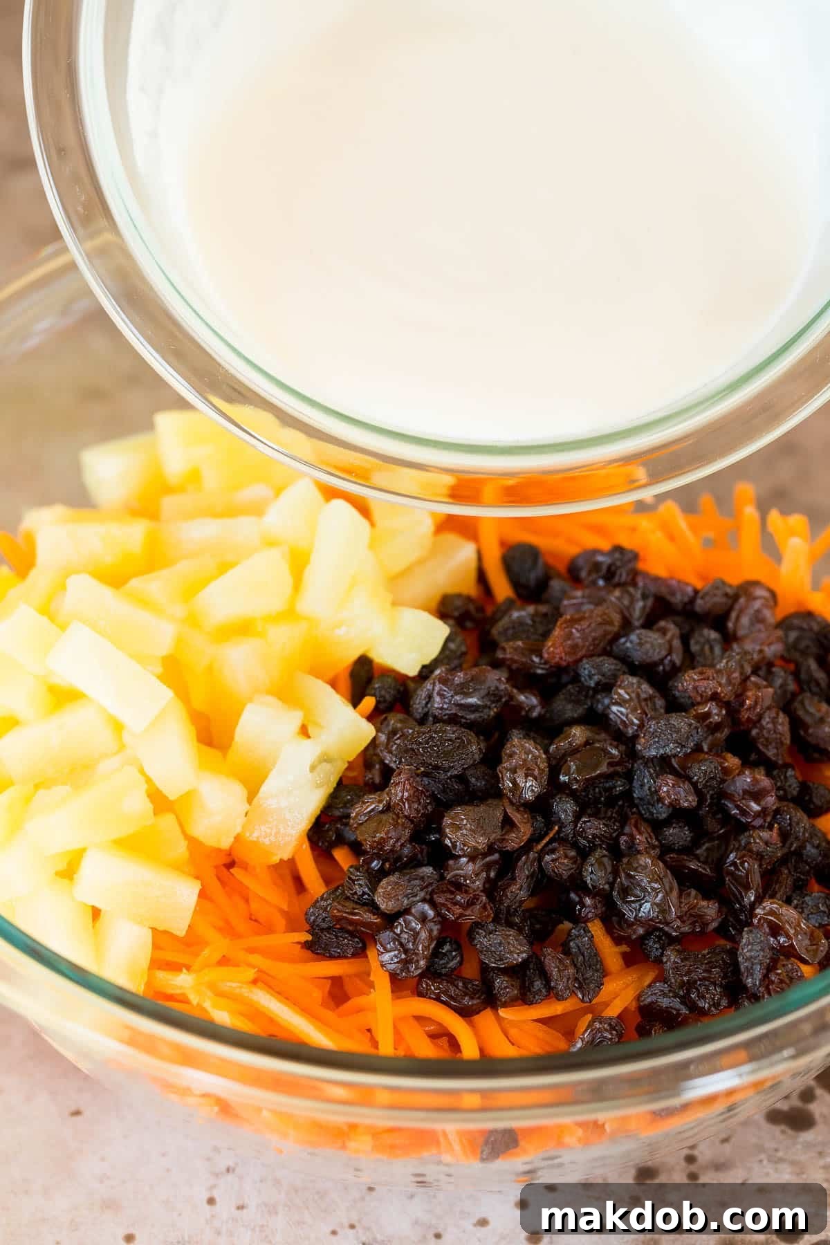 A large serving bowl, with a rich, creamy dressing being poured generously over a colorful mixture of shredded carrots, raisins, and pineapple, ready to be tossed.