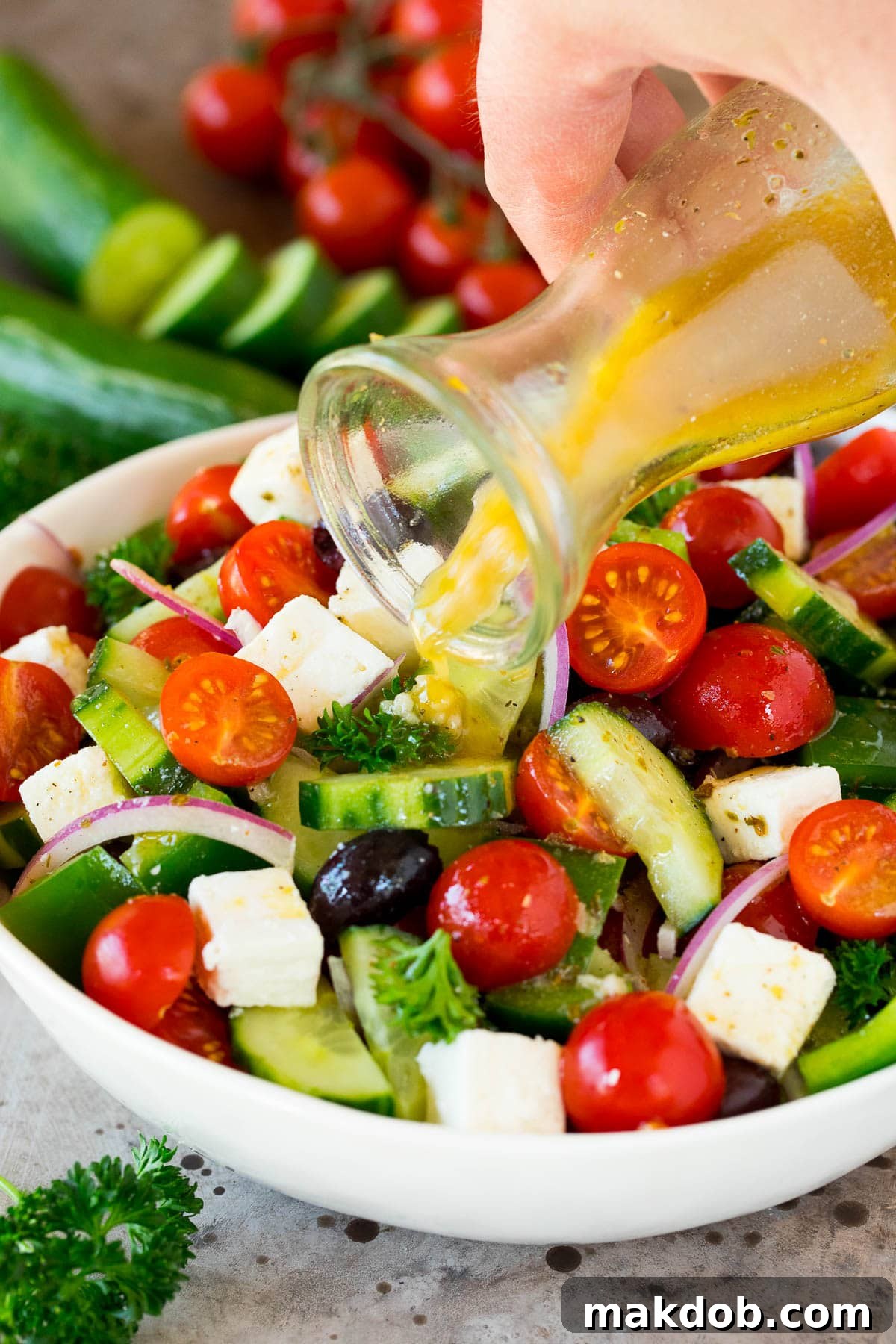 Fresh homemade Greek salad dressing being drizzled generously over a vibrant bowl of diced vegetables, highlighting its rich texture and color.