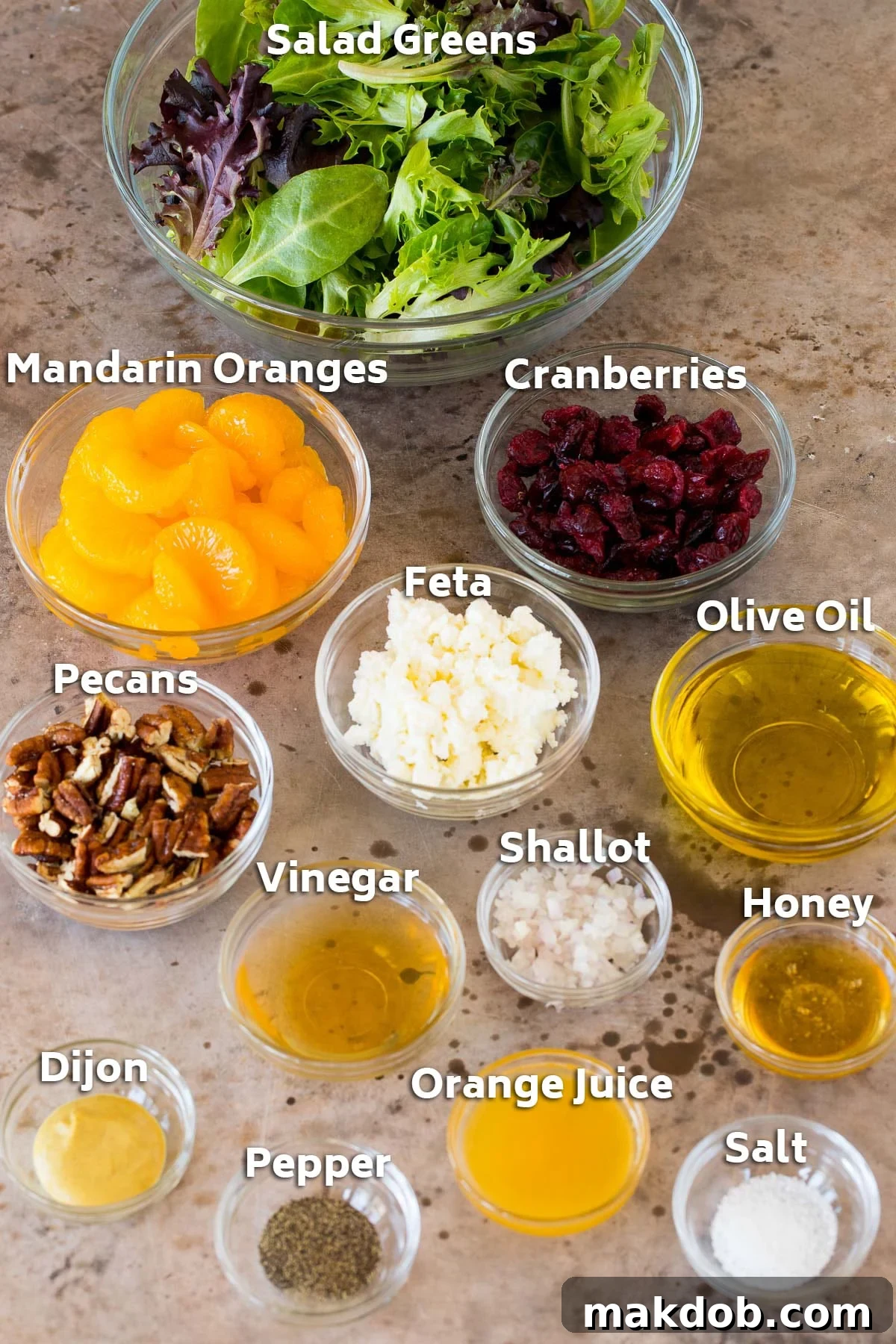 A selection of fresh ingredients laid out, including bowls of mandarin oranges, dried cranberries, candied pecans, crumbled feta cheese, olive oil, apple cider vinegar, honey, Dijon mustard, and a whole shallot.